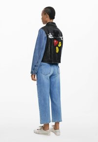 La giacca di jeans e pelle presenta un retro in pelle nera con grafiche di cartoni animati colorate, maniche in denim blu e un design accorciato.