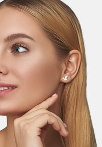 Vita fågelformade örhängen i metall, visade på en modell med rakt blont hår, med fokus på örhängenas design och placering.