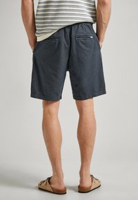 Mężczyzna w ciemnoszarych, casualowych szortach i beżowych sandałach na slip-on, stojący z rękami w kieszeniach na jednolitym tle.