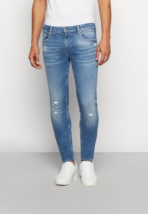 Homme portant un jean slim délavé bleu clair et des baskets blanches, debout devant un fond uni clair.