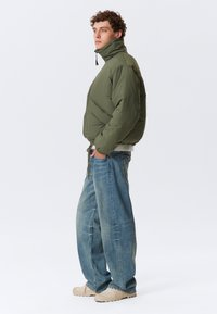 Olivegrøn pufferjakke med høj krave, der har skrå syninger og snøre i kanten, parret med lyseblå vide jeans og beige sko.