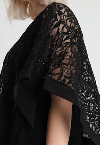 Blonde Person, die ein transparentes schwarzes Spitzenoberteil mit floralen Mustern, Pailletten und weiten Ärmeln trägt, seitlich vor einem schlichten Hintergrund gezeigt.