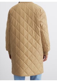 Beżowy, pikowany długi płaszcz z diamentowym przeszyciem, noszony na jasnoniebieskich dżinsach, pokazany z tyłu na stojącej osobie.