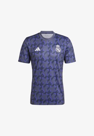 Homme portant un maillot de football Real Madrid bleu et violet à motifs et un pantalon Adidas bleu foncé, debout devant un fond uni.