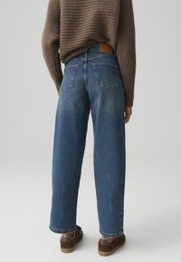 Wide-leg blue denim jeans with a faded wash, two back pockets, and a brown leather patch on the waistband, paired with brown shoes.
