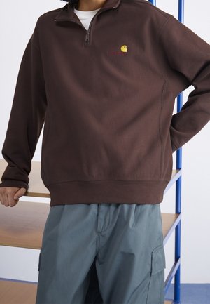 Persona vistiendo un sudadera Carhartt marrón con cremallera hasta el pecho y pantalones cargo azul claro, apoyada en una mesa de madera con un marco metálico azul.