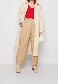 Manteau trench crème, haut tricoté rouge et pantalons larges beiges. Les détails incluent des boutons sur le manteau et un sac à main blanc. Bottes à talon bloc tan.