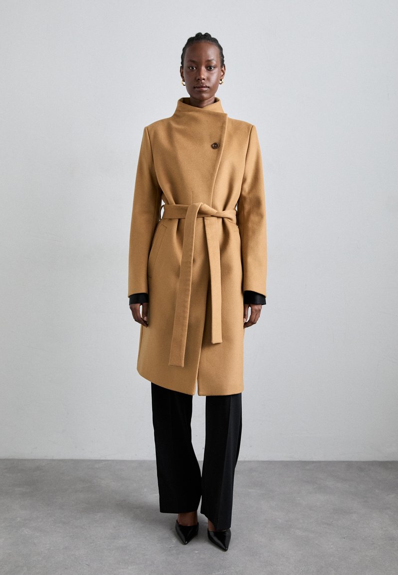 Camel wool coat with high collar, single button closure, belt at waist, and black cuff accents, paired with black pants and pointed heels.