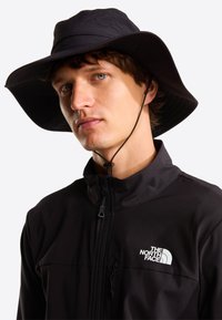 Μαύρο καπέλο με ευρύ γείσο και υφή υφάσματος, με λουράκι κάτω από το πηγούνι για σταθερή εφαρμογή. Στο μπροστινό μέρος εμφανίζεται το λογότυπο της The North Face.