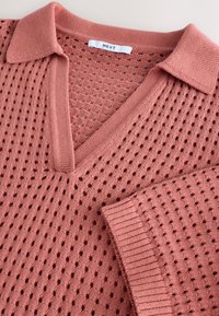 Megztinis iš purvinos rožinės spalvos siūlų, turintis V formos iškirptę ir apykaklę. Dizainą puošia atvirų akių raštai ir ribbinga rankogalių apdaila, suteikianti tekstūrą ir detales.