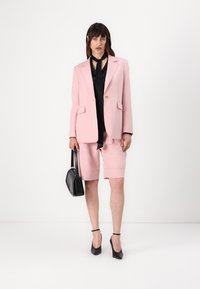 Blazer rosa chiaro con tasche, abbinato a pantaloni corti coordinati. Blusa nera sotto, accessoriata con una borsa nera e tacchi.