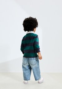 Rohelise ja tumesinise triibuline pika varrukaga särk, valge krae, sobitatud lahtiste tuhmunud teksapükstega ja valgete vabaajajalatsitega. Laps nähtav tagantpoolt.