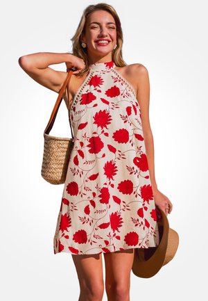 Lächelnde Frau in einem ärmellosen weißen Kleid mit rotem Blumenmuster, die eine gewebte Tasche über der Schulter trägt und einen Strohhut in der Hand hält.