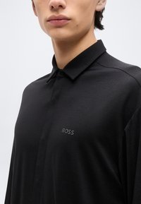 Κοντινό πλάνο ενός ατόμου που φοράει μαύρο πουκάμισο με γιακά, με κρυφή σειρά κουμπιών και διακριτικό λογότυπο «BOSS» στο στήθος.