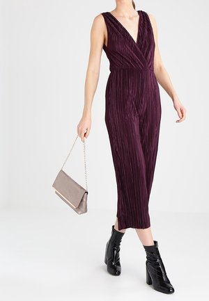 Bezruký švestkový plisovaný overal s výstřihem do V, nošený s černými lakovanými kotníkovými botami, držící béžovou kabelku s řetízkovým popruhem.