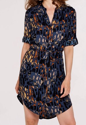 Vrouw die een overhemdjurk met korte mouwen draagt in marineblauw, oranje en beige met een strik in de taille en opgerolde manchetten.