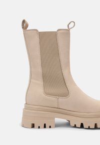 Béžová kotníková bota s hladkým koženým vnějškem a elastickými bočními panely. Má chunky texturovanou podrážku a poutko pro snadné obutí.