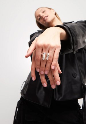 Anillos de plata con un diseño liso y alargado en los dedos, mostrados sobre un fondo claro, manos parcialmente extendidas, destacando los anillos.