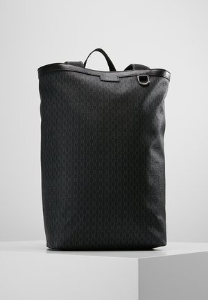 Wysoki czarny plecak z teksturowanej tkaniny z subtelnym wzorem, skórzanymi wykończeniami, górnym uchwytem, pierścieniem D i wyściełanymi paskami na ramiona na białym piedestale.