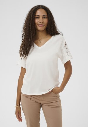 T-shirt blanc à col en V avec manches courtes ornées de dentelle, associée à un pantalon marron clair. Texture lisse, coupe décontractée. Collier doré visible.
