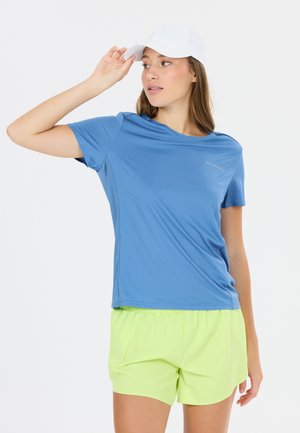 Ung kvinde iført en hvid kasket, blå kortærmet skjorte og neongule shorts, der kigger til siden med hånden, der rører kasketten.