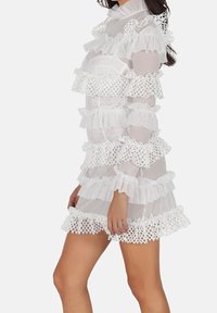Robe blanche et transparente dotée de couches froncées avec des accents en dentelle, des manches longues et un col haut, offrant un aspect léger et texturé.