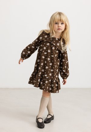 Jeune fille aux longs cheveux blonds portant une robe fleurie marron, des chaussettes beiges montantes et des chaussures Mary Jane noires, debout sur un sol clair uni.