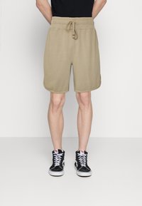 Pantalones cortos deportivos color caqui con cintura elástica y cordón. Presenta bolsillos laterales y una textura suave. Combinados con zapatillas altas negras.