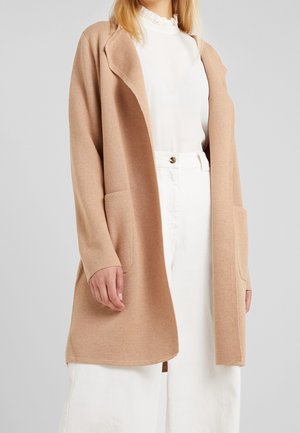 Frau trägt eine beige Strickjacke mit offenem Vorderteil über einer weißen Bluse mit gerafftem Kragen und hoch taillierter weißer Hose vor einem einfarbigen Hintergrund.