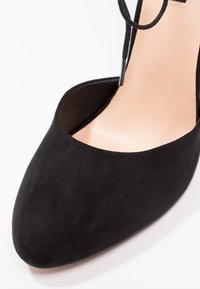 Chaussure à talon haut en suede noir avec un bout pointu et des côtés ouverts, dotée d'un fin détail en dentelle noire près de la cheville. Doublure intérieure lisse.