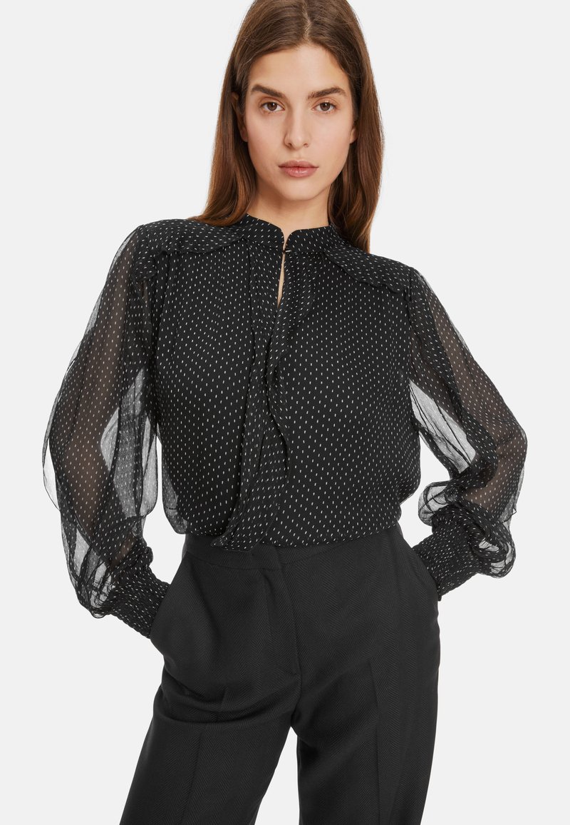 Schwarze Bluse mit Polka-Dots aus durchsichtigem Material, mit langen Puffärmeln und einem geknöpften Kragen, kombiniert mit hochtaillierten schwarzen Hosen.