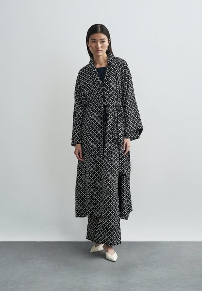 Μαύρη μακριά ρόμπα με λευκό γεωμετρικό σχέδιο, ζωνάτη στη μέση, με φαρδιά μανίκια και ρευστή σχεδίαση, φορεμένη πάνω από μαύρο φόρεμα.