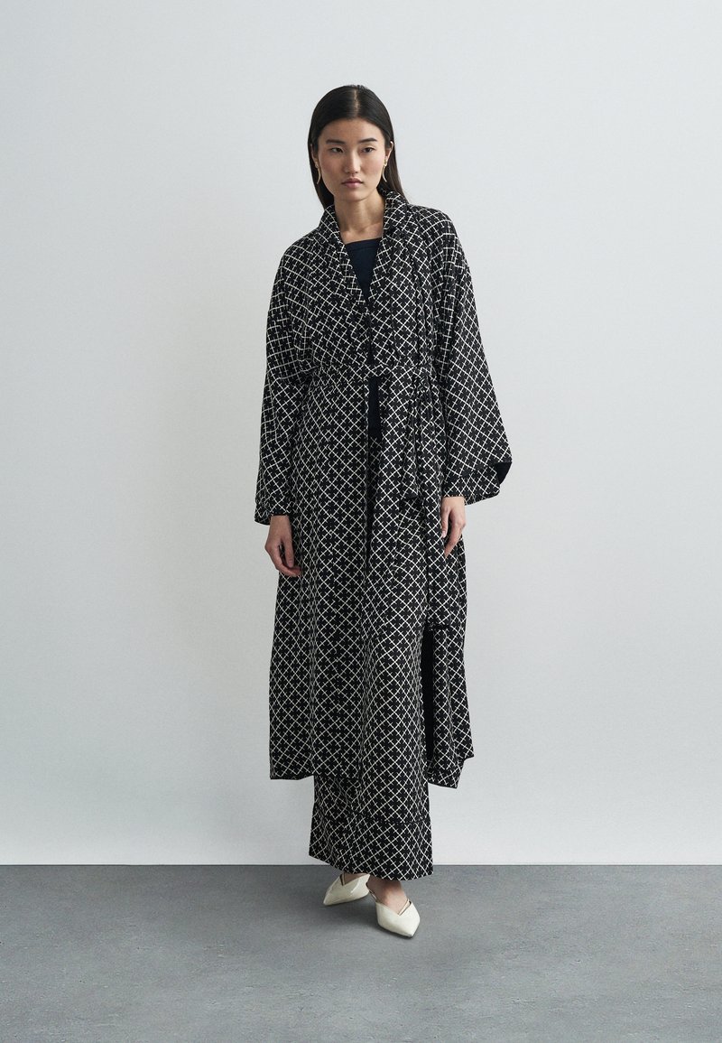 Langes schwarzes Gewand mit einem weißen geometrischen Muster, tailliert in der Taille, mit weiten Ärmeln und einem fließenden Design, getragen über einem schwarzen Kleid.