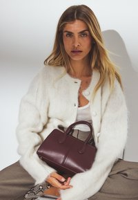 Djup burgunder läderhandväska med korta handtag, slät yta och kontrasterande sömmar. Kvinna i en fluffig vit cardigan och mönstrade byxor.