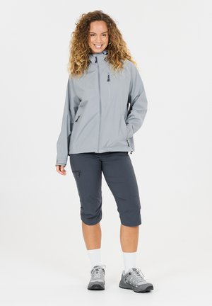 Lächelnde Frau mit lockigem Haar, die eine hellgraue Jacke, dunkelgraue gekürzte Hosen, weiße Socken und graue Wanderschuhe trägt und vor weißem Hintergrund steht.