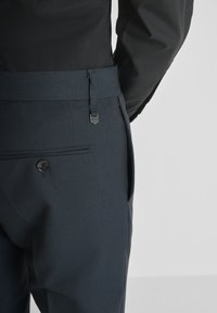 Man in een donkere, gestructureerde broek met een knoopzak achter en een brede bijpassende riem, gecombineerd met een zwart overhemd met lange mouwen.
