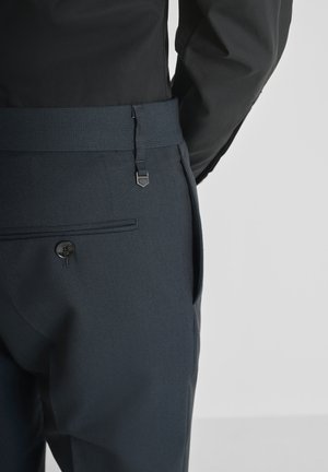 Uomo che indossa pantaloni scuri con una texture, dotati di una tasca posteriore con bottone e una cintura larga abbinata, abbinati a una camicia nera a maniche lunghe.