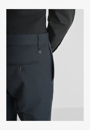 Uomo che indossa pantaloni scuri con una texture, dotati di una tasca posteriore con bottone e una cintura larga abbinata, abbinati a una camicia nera a maniche lunghe.