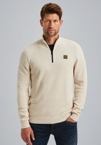 Crème-kleurige pullover met een kwart-rits kraag, gestructureerde stof en ribgebreide manchetten, met een klein, gemarkeerd label op de borst.