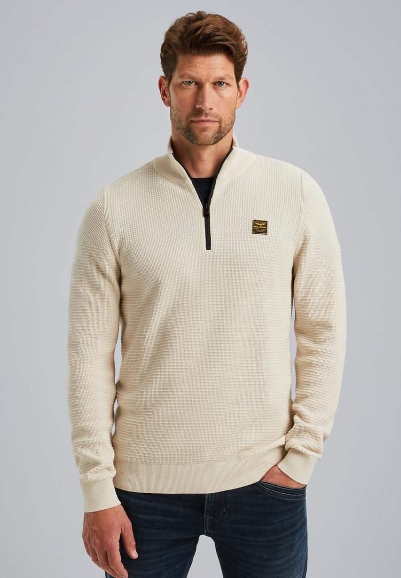Crème-kleurige pullover met een kwart-rits kraag, gestructureerde stof en ribgebreide manchetten, met een klein, gemarkeerd label op de borst.