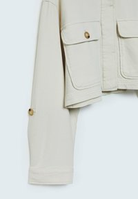 Manche de veste beige clair avec poche à rabat boutonnée et détail de bouton au poignet, présentée sur un fond clair uni.