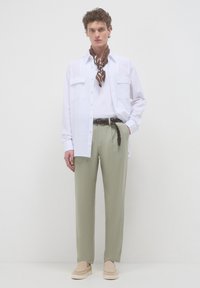 Camicia bianca a maniche lunghe, pantaloni verdi, sciarpa marrone a motivi legata al collo e espadrillas beige si stagliano su uno sfondo chiaro.