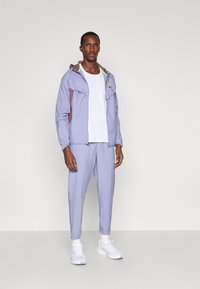 Άνδρας φορώντας ανοιχτό μοβ φόρμα και παντελόνι πάνω από λευκή μπλούζα και λευκά αθλητικά παπούτσια, στέκεται σε απλό φόντο.