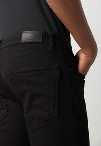 Czarne jeansy z denimu o gładkiej fakturze, z tylnymi kieszeniami i skórzaną naszywką z logo na pasie. Prosty design z minimalnymi detalami.