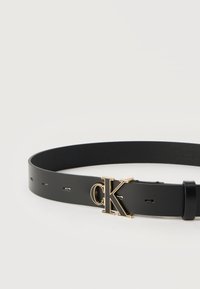 Μαύρη δερμάτινη ζώνη με αγκράφα λογότυπο "cK" σε χρυσό τόνο και πολλαπλές τρύπες ρύθμισης, εμφανιζόμενη σε λευκό φόντο.