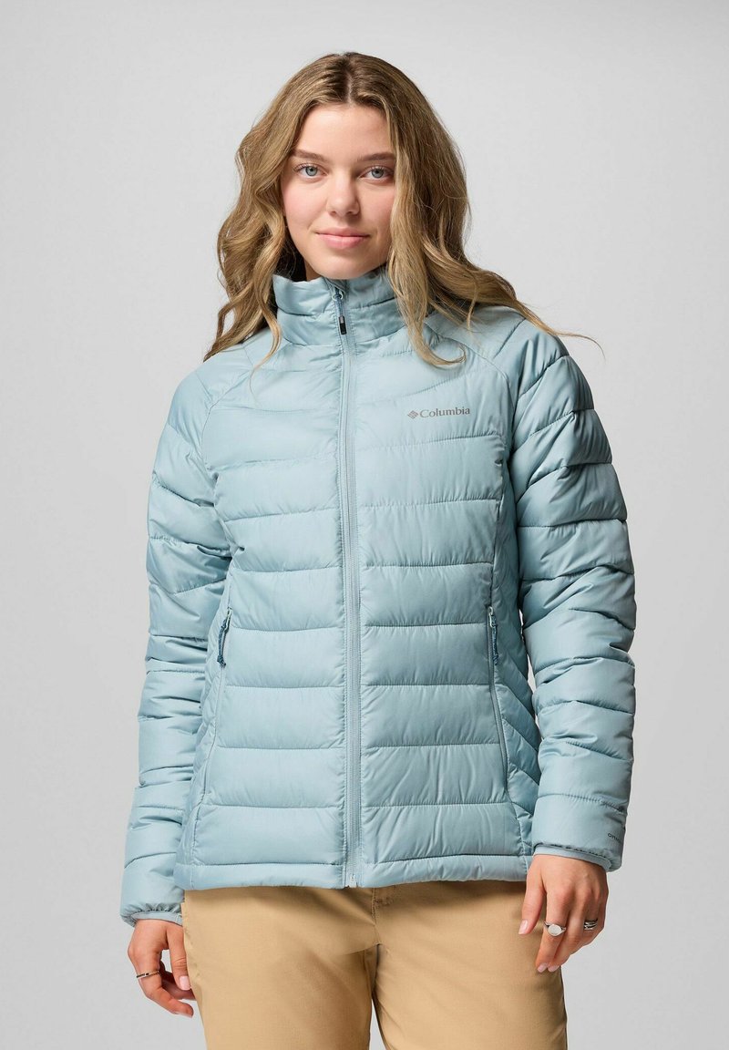Frau mit langen, gewellten Haaren, die eine hellblaue gesteppten Columbia-Jacke und beigen Hosen trägt und vor einem schlichten grauen Hintergrund steht.