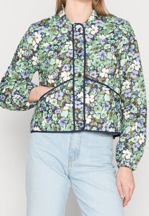 Vrouw die een bloemige doorgestikte jas draagt met drukknoopsluiting, donkere bies en elastische manchetten, gecombineerd met lichtblauwe, hooggetailleerde jeans.