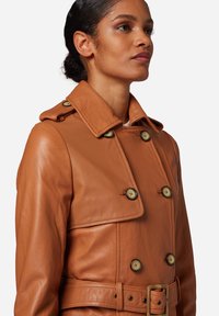 Brown leather trench coat with a double-breasted design, large buttons, and a belted waist. Features a wide collar and smooth texture.