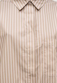 Μπεζ και άσπρο πουκάμισο με κάθετες ρίγες, κουμπιά, μυτερό γιακά και τσέπη στο στήθος στη δεξιά πλευρά.