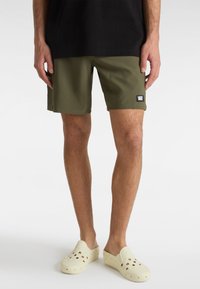 Shorts vert olive en tissu léger, arborant un écusson logo Vans, avec une coupe décontractée et droite. Portés avec des sabots beiges.